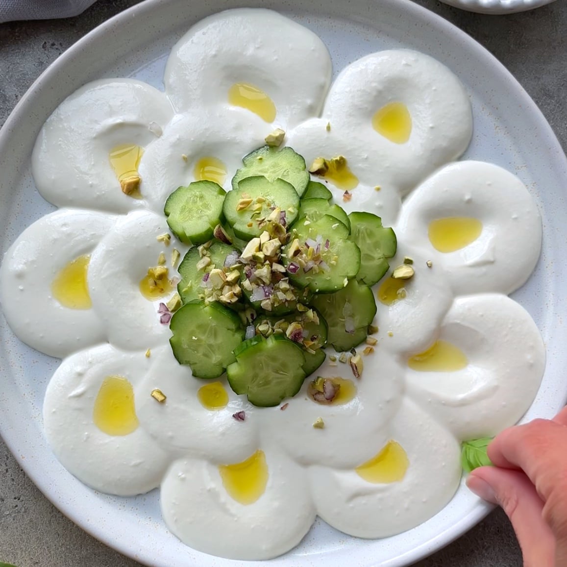 A plate of whipped feta dip spread in circles, topped with sliced cucumbers, chopped nuts, red onion, and drizzled with olive oil. A hand is placing a basil leaf on the edge.