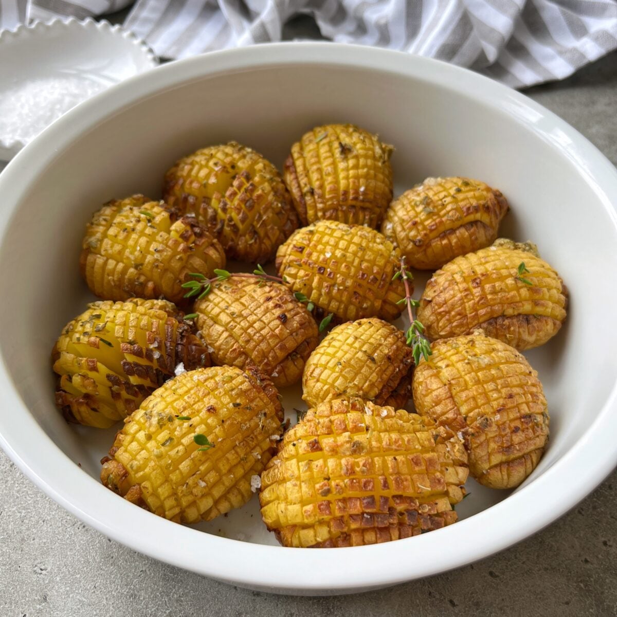 A white bowl filled with several Hasselback Baked Potatoes, garnished with herbs and coarse salt, sits on a gray surface.