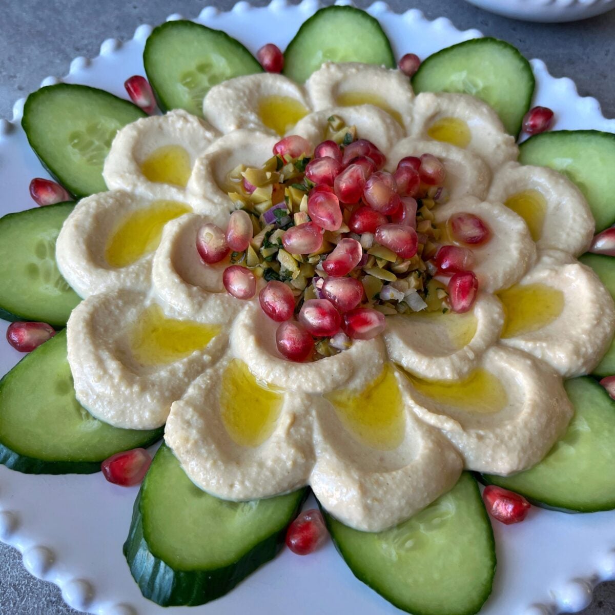 A Hummus Platter garnished with olive oil, pomegranate seeds, and chopped pistachios, surrounded by cucumber slices on a white plate.