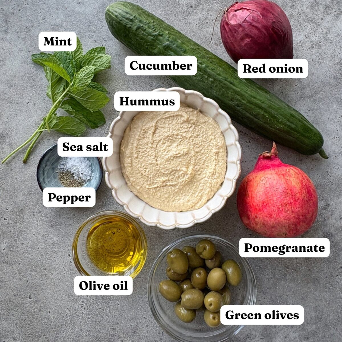 Ingredients arranged on a gray surface including olives, a cucumber, red onion, and hummus.