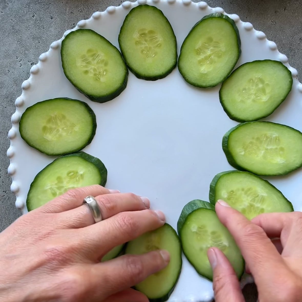 Hands arrange slices of cucumber along the outer edge of a white platter.