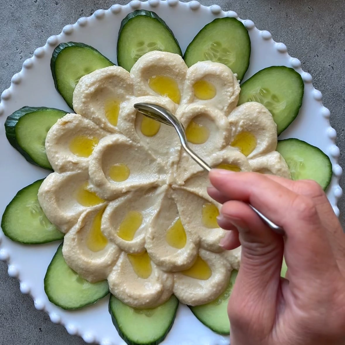 A hand spoons olive oil in the center of each hummus petal.