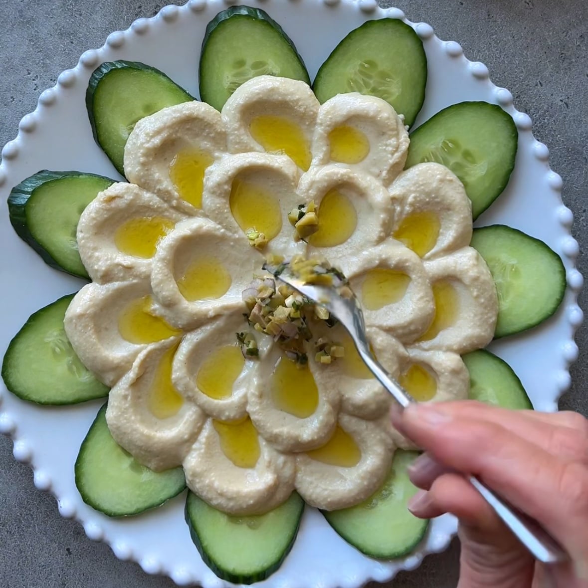 A hand spoons olives in the center of a hummus platter.