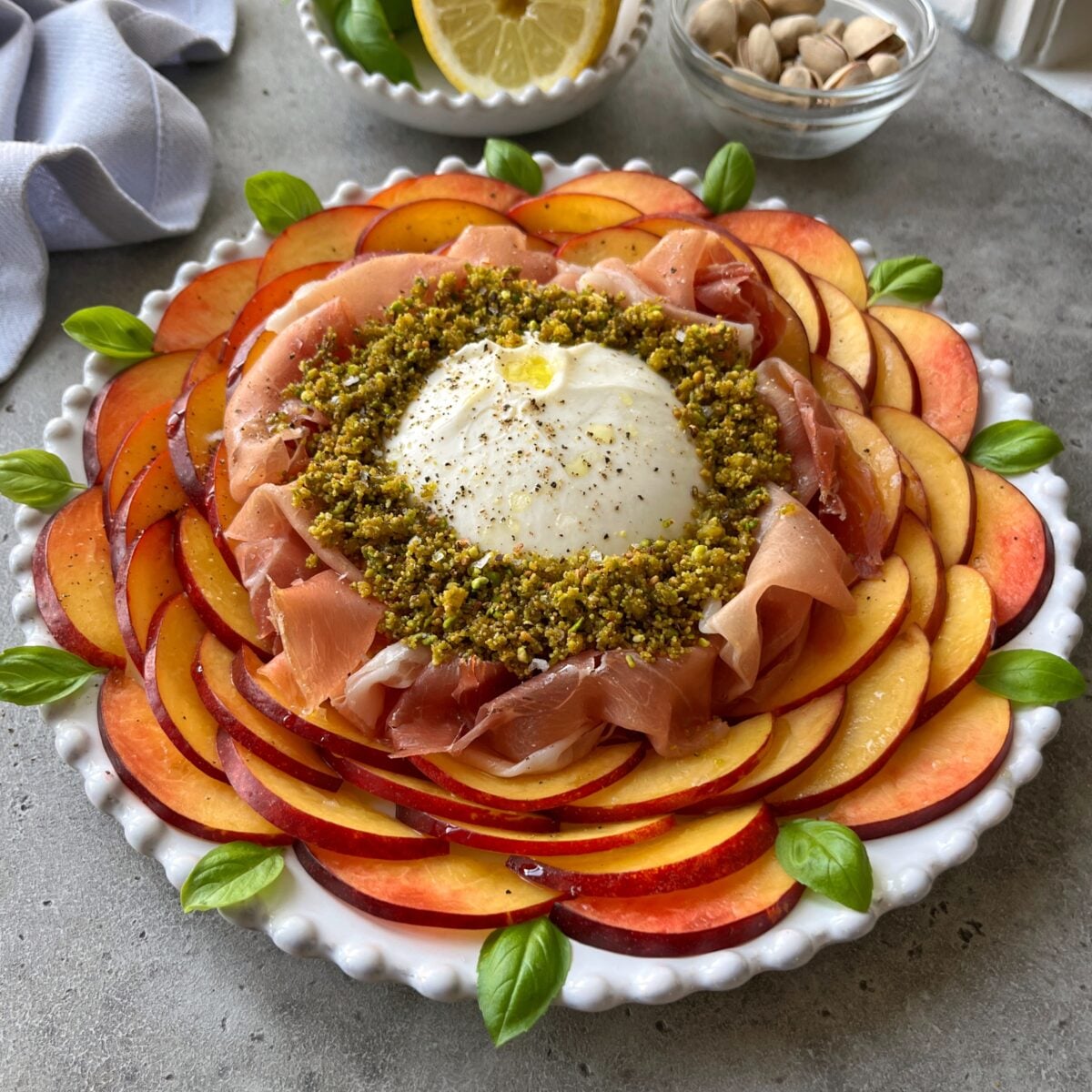 A plate with sliced nectarine arranged in a circle, topped with Serrano, burrata cheese, crushed pistachios, basil leaves, and a lemon wedge in the background.