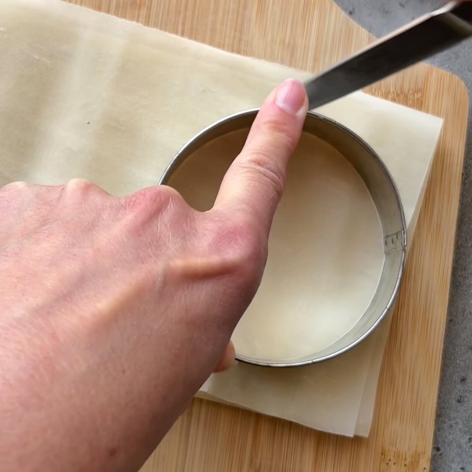 A hand holds a round metal cutter on top of layered pastry sheets, preparing to cut a circle on a wooden board.