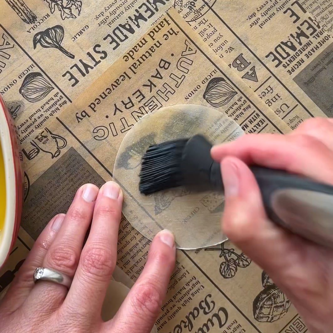 A hand uses a silicone brush to apply melted butter to a round sheet of dough on top of printed parchment paper.