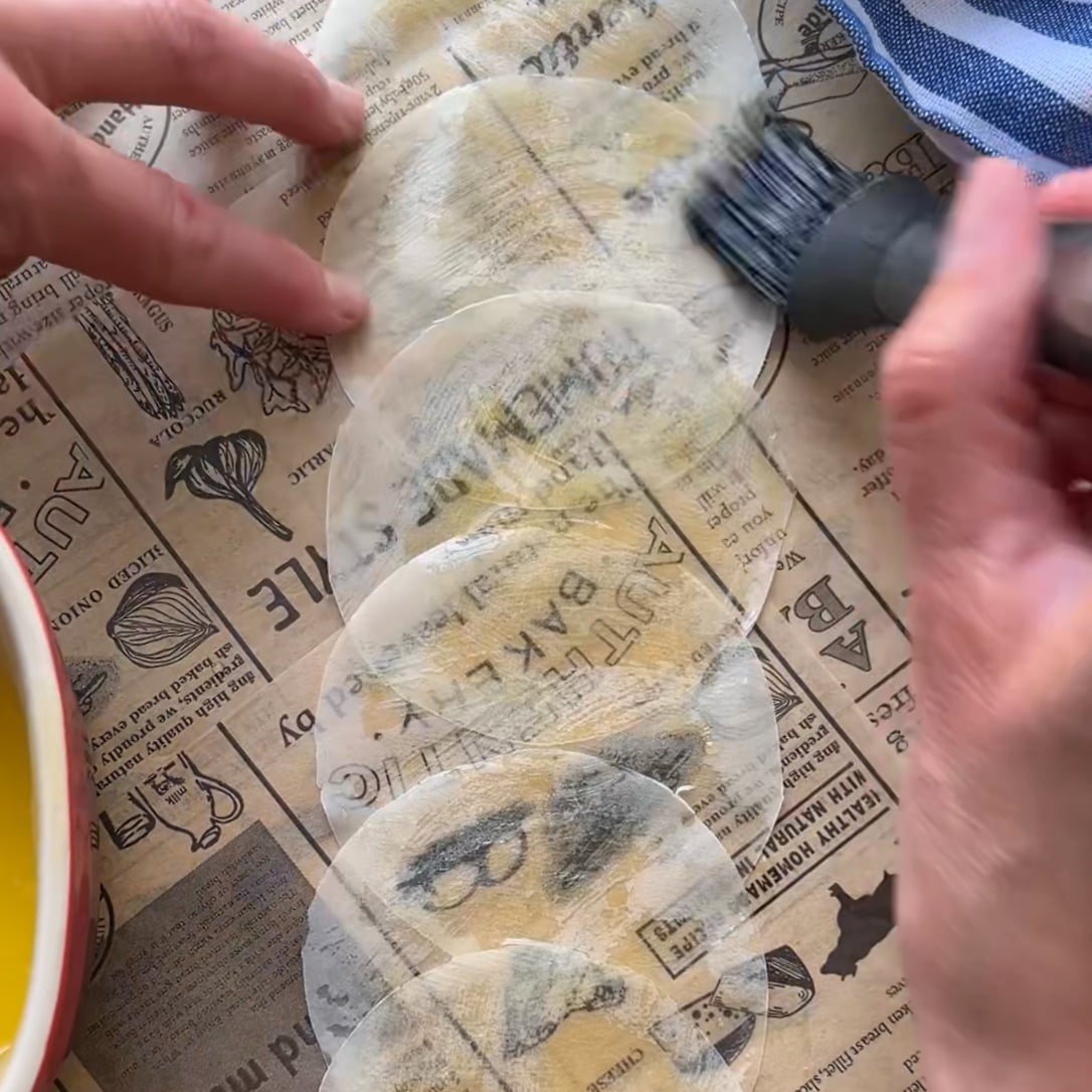 A person brushes melted butter onto a row of round phyllo wrappers placed on printed parchment with a bowl of melted butter and a striped cloth nearby.