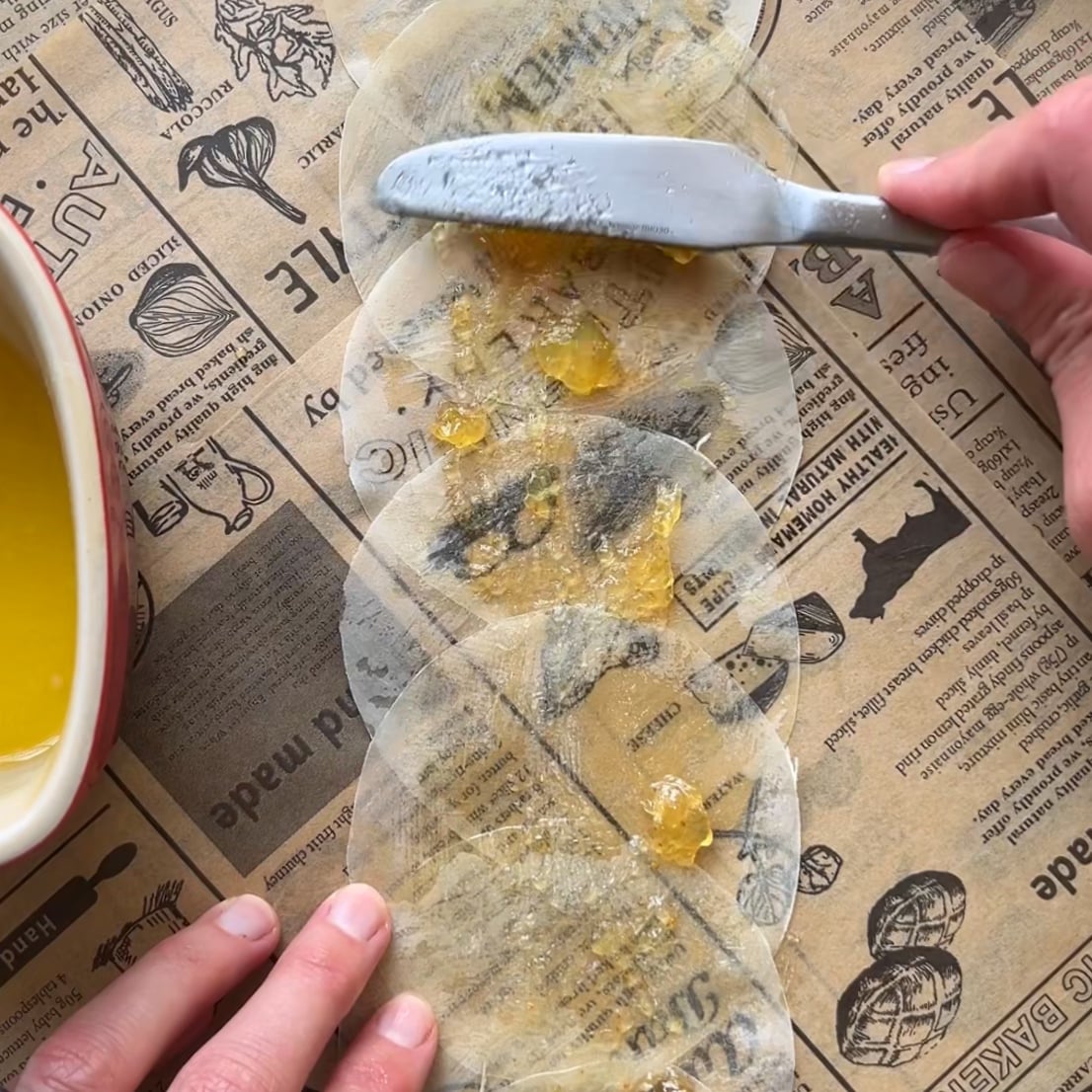 A person spreads a yellow fig jam onto overlapping phyllo paper circles laid on printed parchment paper, using a knife.