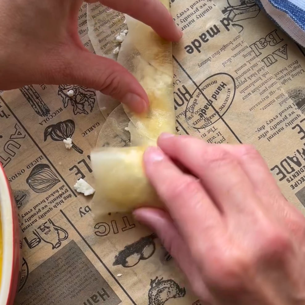 Hands rolling a phyllo dough appetizer into a rose shape on printed parchment paper.