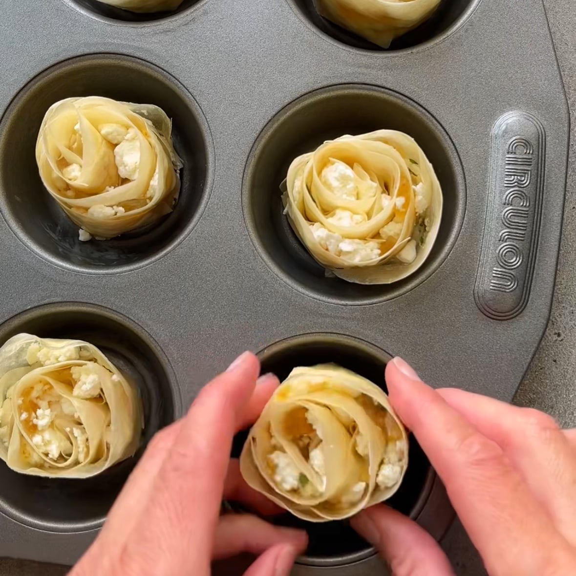 Hands placing a rolled phyllo dough appetizer with cheese filling into a muffin tin, with five other similar phyllo dough appetizers already in the tin.