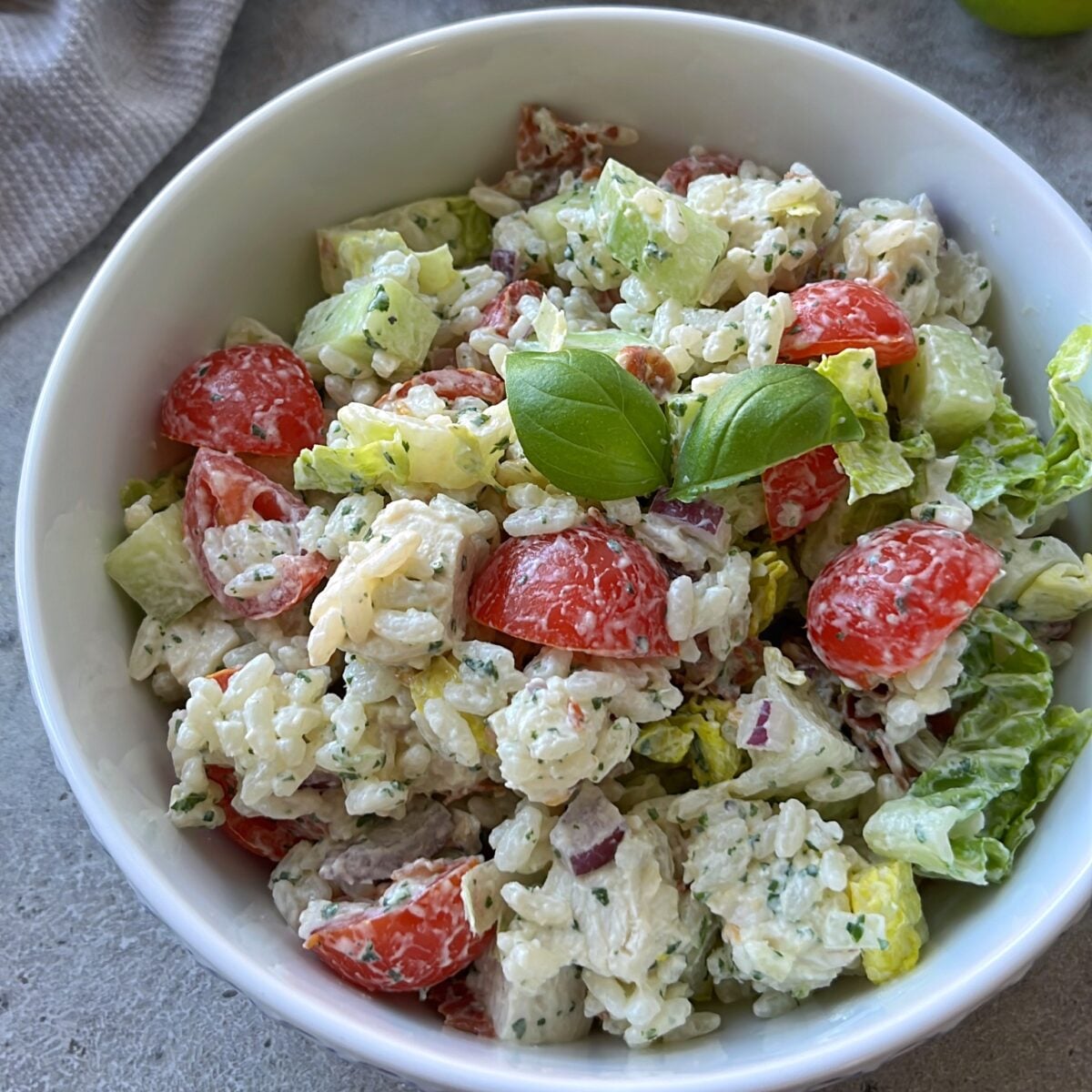 A bowl of creamy Rice Chicken Salad with cherry tomatoes, lettuce, red onion, cucumber, and a fresh basil garnish.