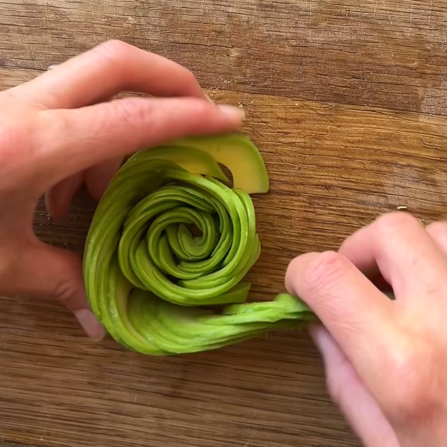 Hands arranging thin slices of avocado into a rose shape on a wooden cutting board.