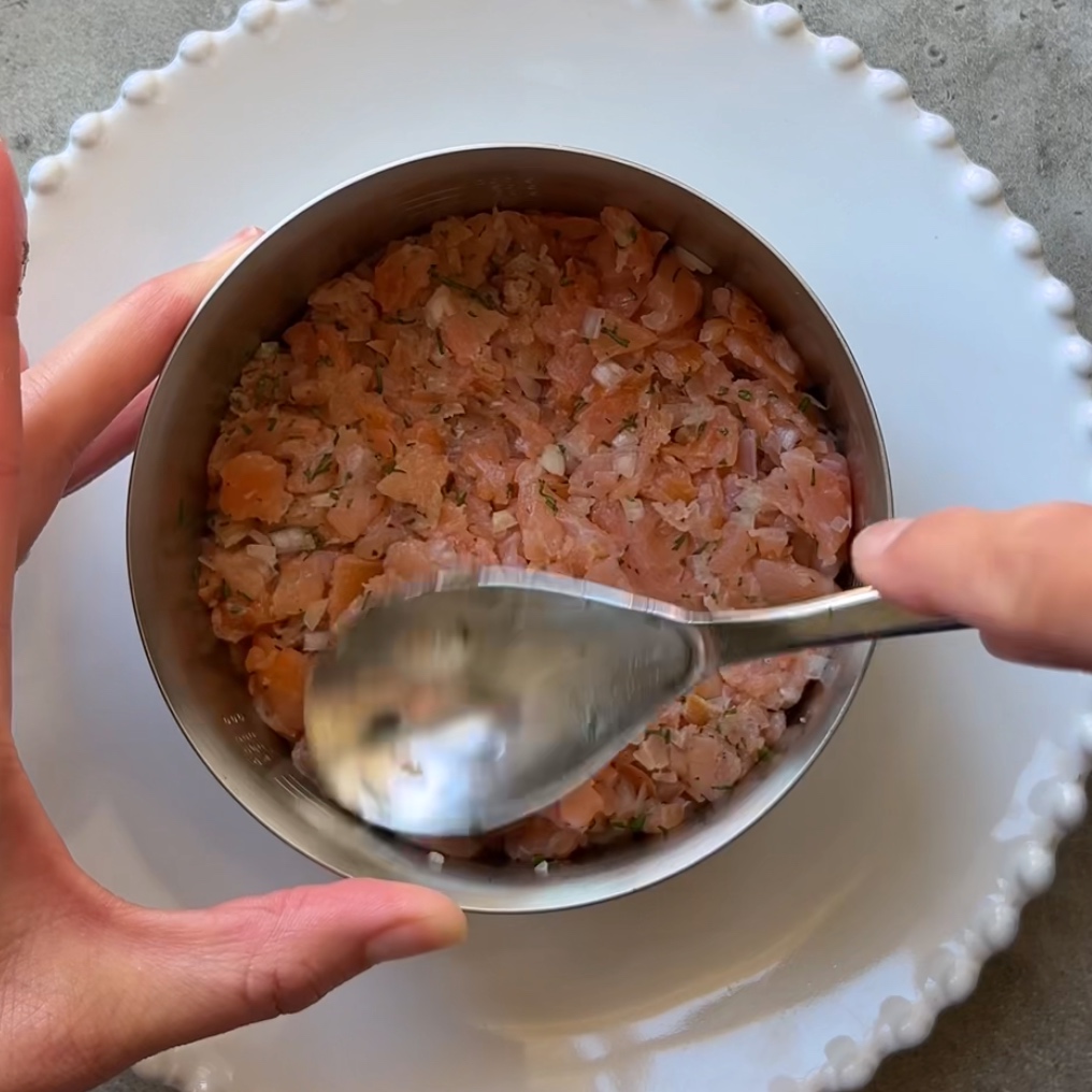A person presses chopped raw salmon into a metal ring mold on a white plate using a spoon.