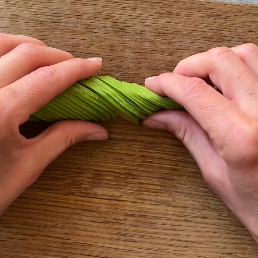 Two hands arrange thinly sliced avocado on a wooden cutting board, creating the perfect base for a delicious Smoked Salmon Tartare.
