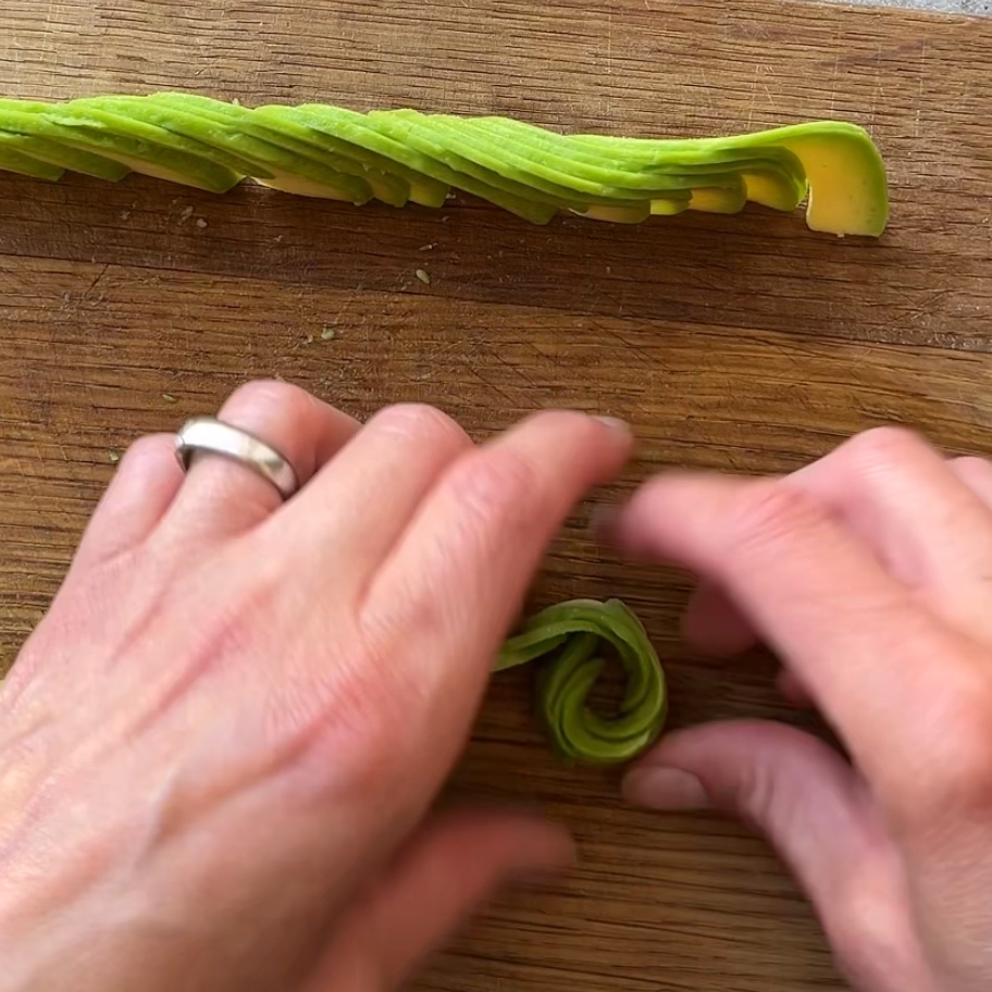 Two hands shape thin avocado slices into a rose on a wooden cutting board. A stack of sliced avocado is in the background.