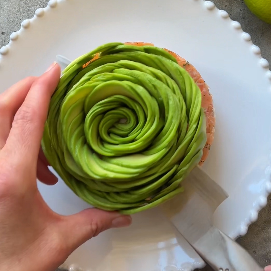 A hand places a spiral arrangement of thinly sliced avocado on top of a Smoked Salmon Tartare beautifully presented on a white plate with a scalloped edge.
