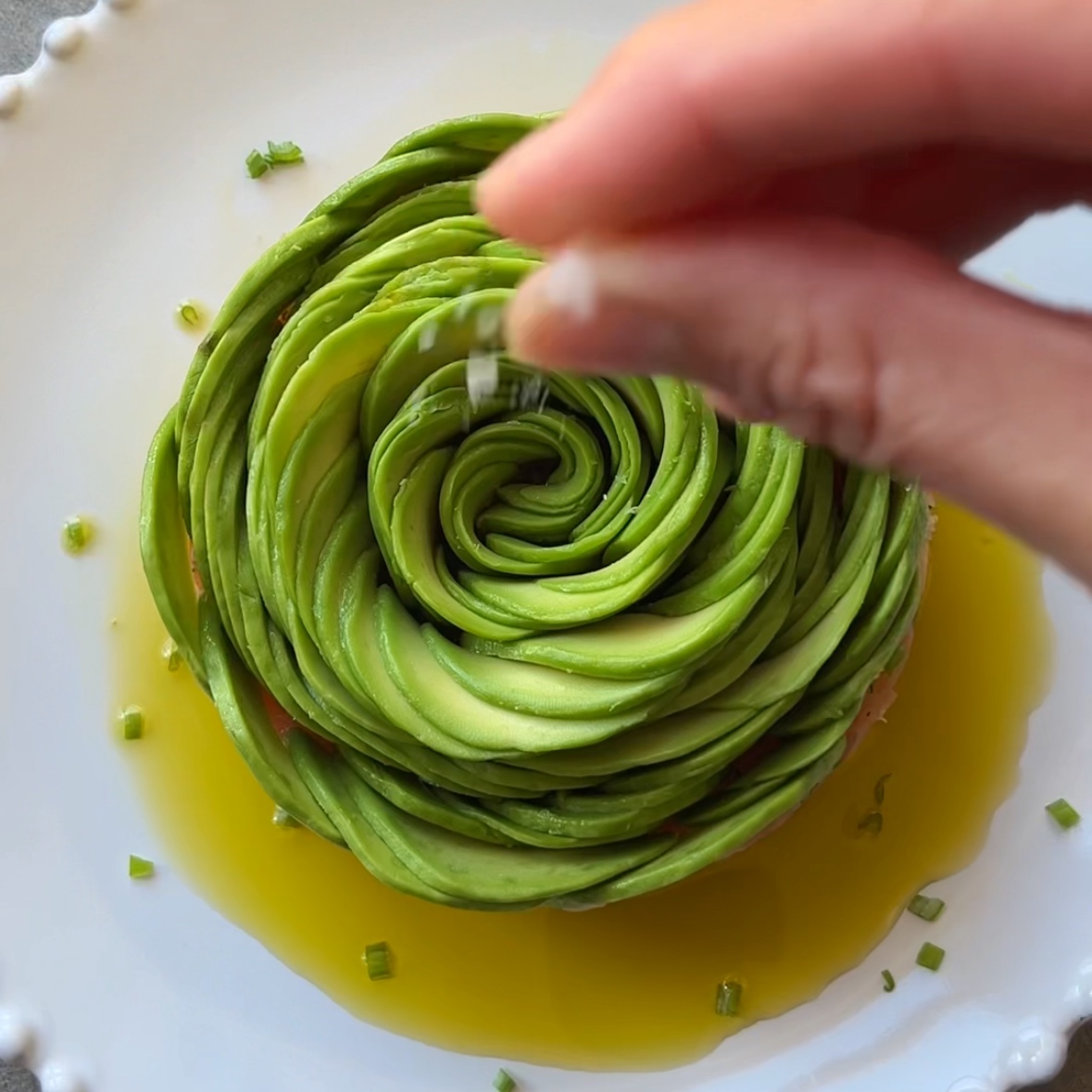 A hand sprinkles salt over an avocado rose arranged on a white plate, surrounded by olive oil and garnished with chopped chives.