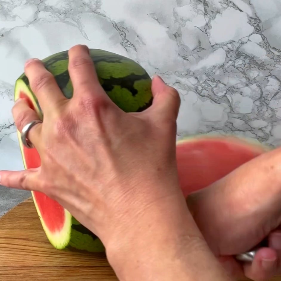 A person slices the top and bottom of a watermelon on a wooden surface using a knife.