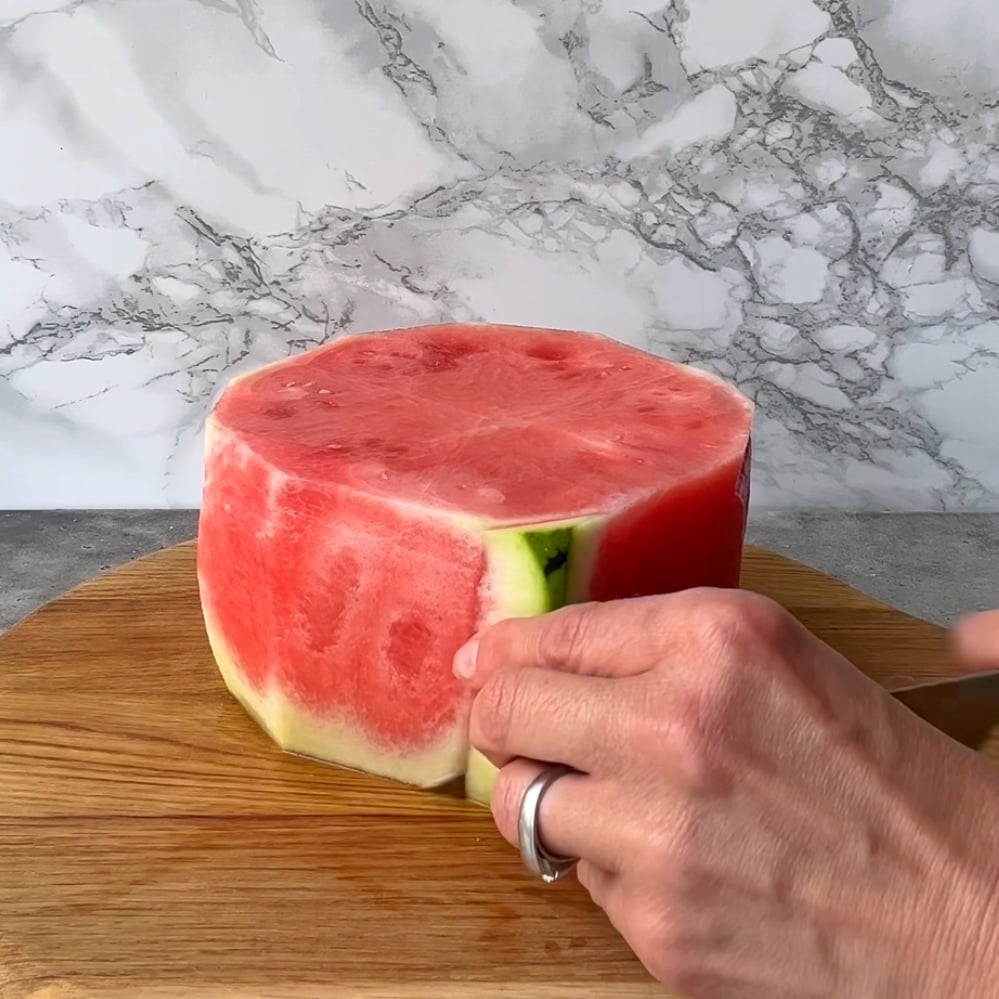 A person slices the rind off a large, round piece of watermelon on a wooden cutting board.