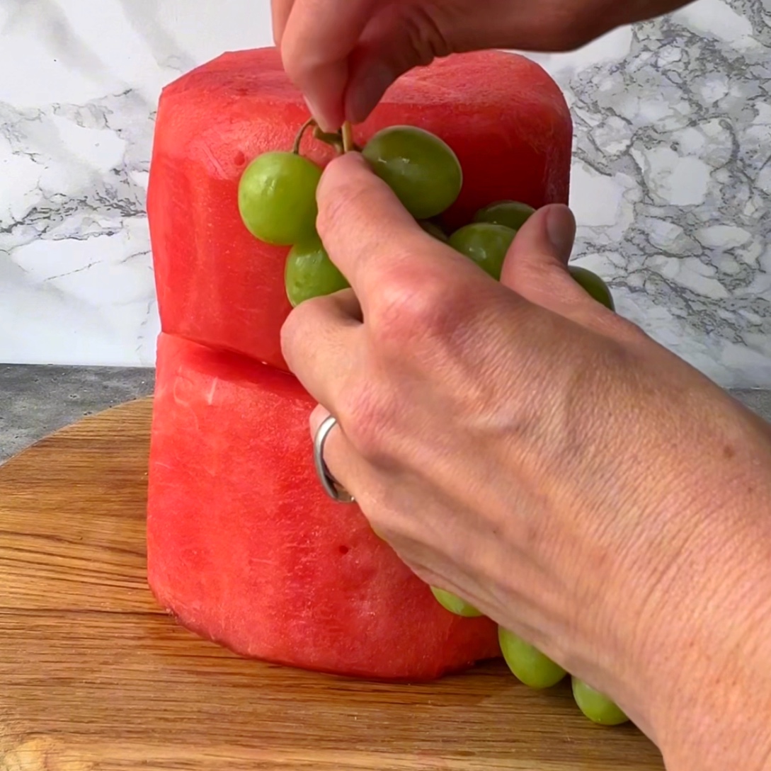 Two hands attach a bunch of green grapes to a watermelon cake.