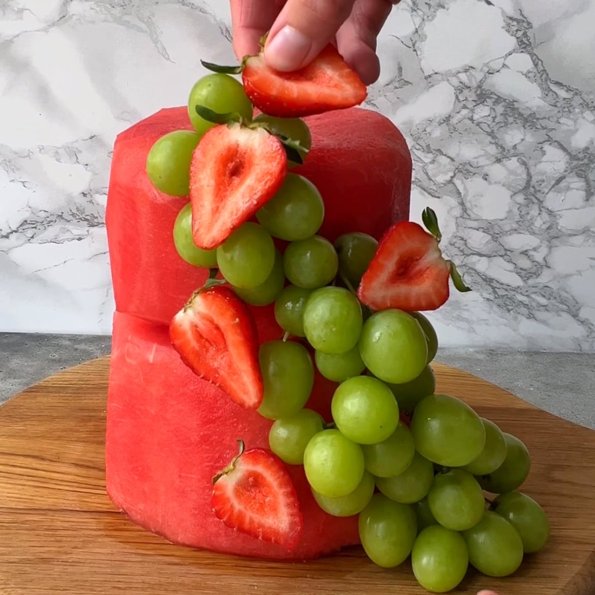 A hand places strawberries on two stacked, peeled watermelon rounds.