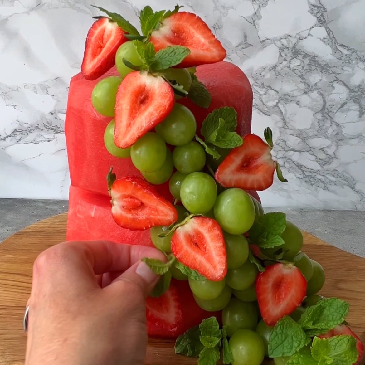 A hand places mint leaves on a two-tiered Watermelon Fruit Cake.
