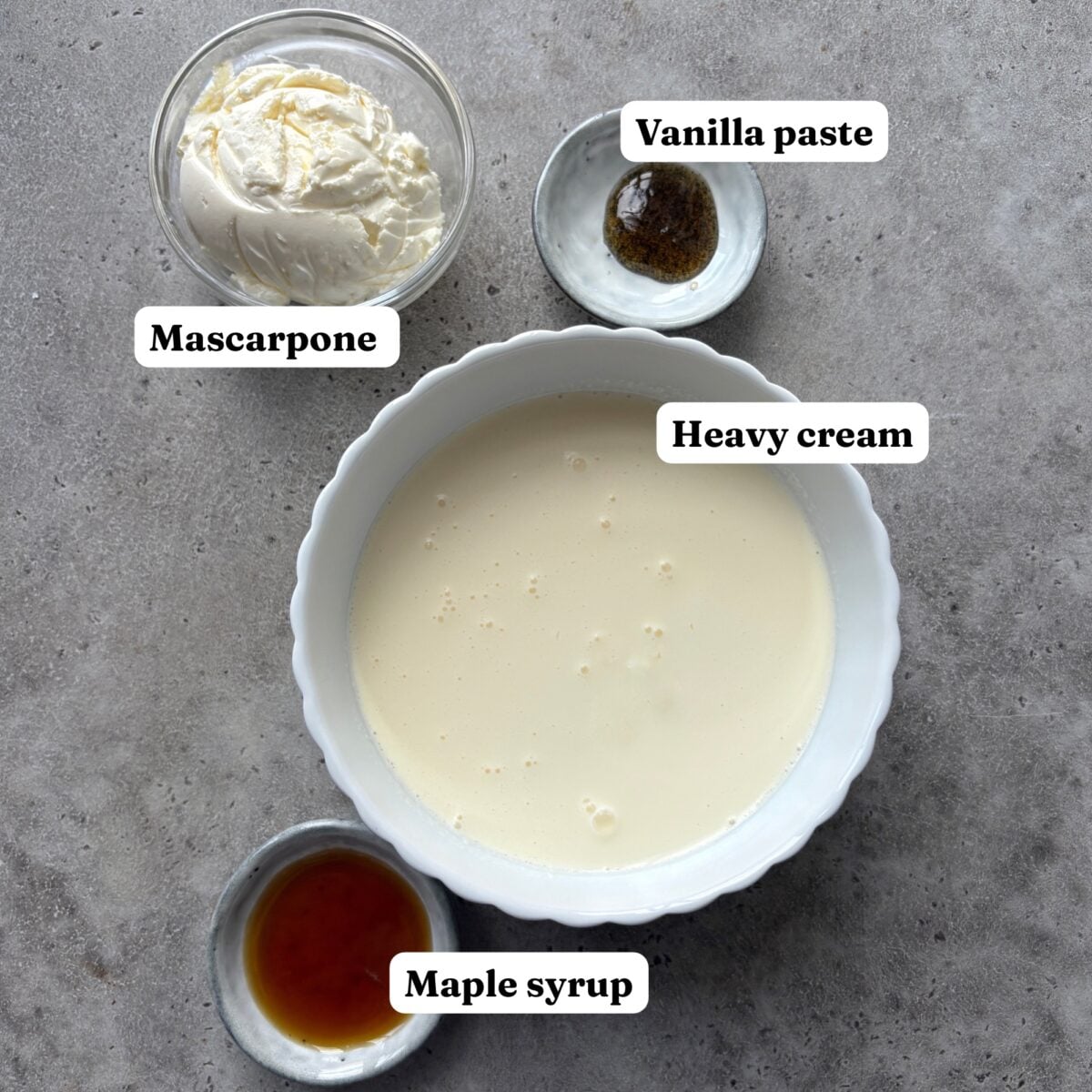 A bowl of heavy cream, a dollop of mascarpone, vanilla paste, and a small dish of maple syrup are arranged on a gray surface, each labeled.