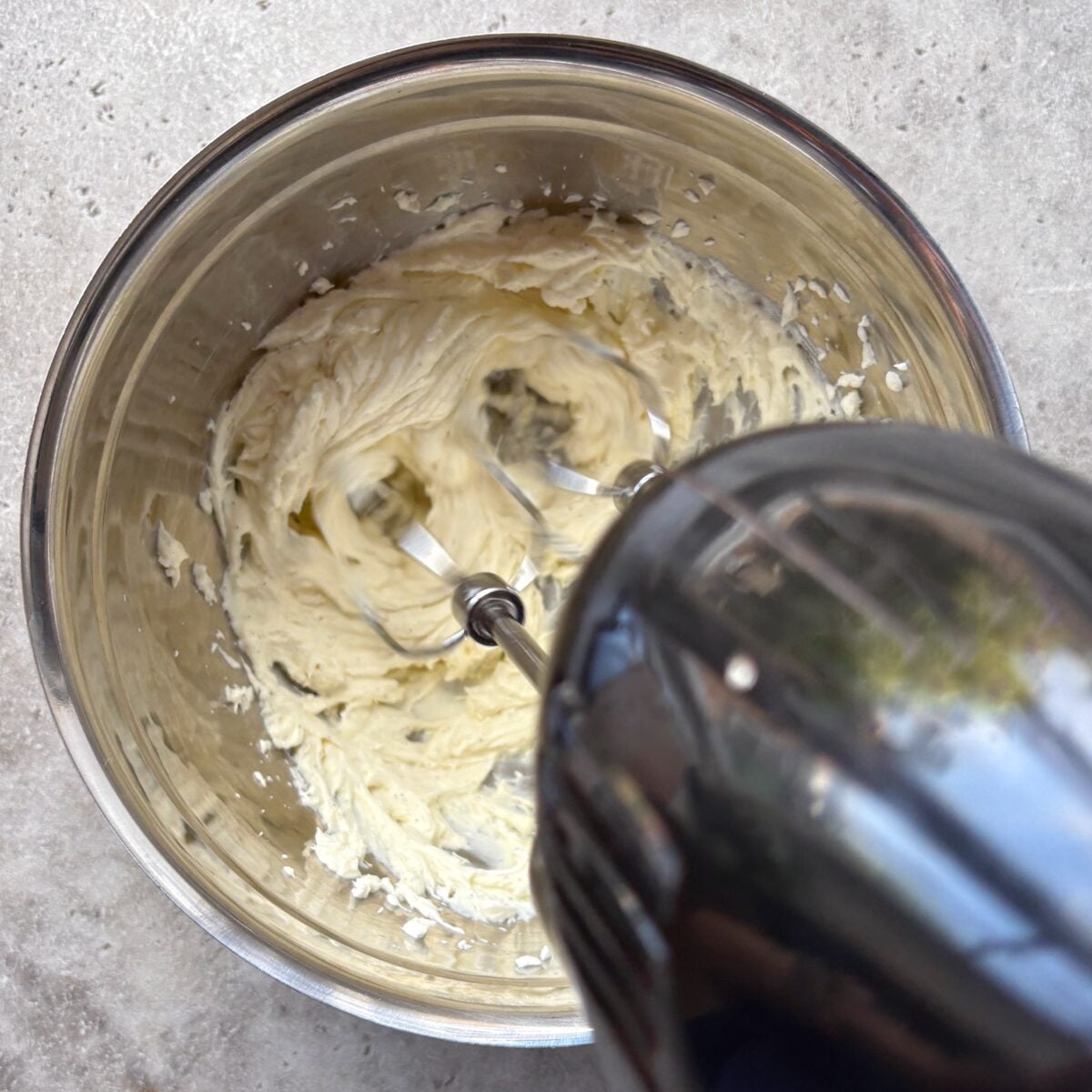 An electric hand mixer blends mascarpone and vanilla paste in a metal bowl on a gray countertop.