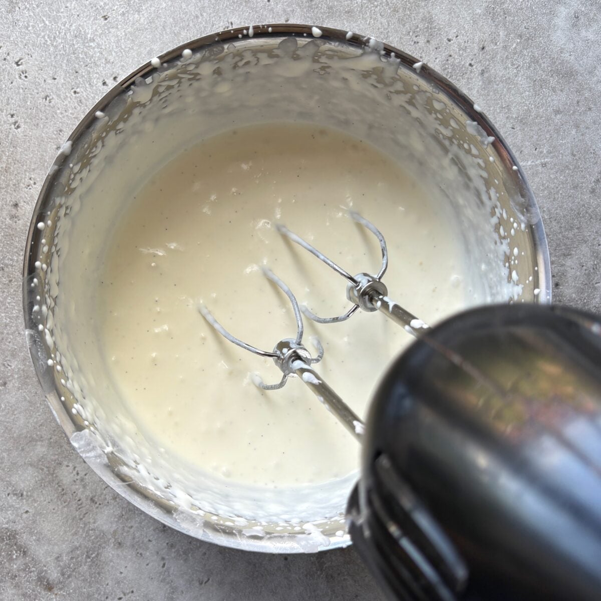 A hand mixer with two beaters is mixing a bowl of creamy 4th of July Dessert Dip batter on a grey countertop.