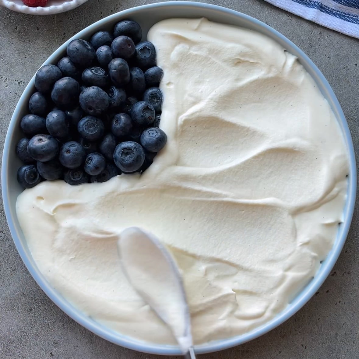 A bowl with creamy white 4th of July Dessert Dip topped with a cluster of fresh blueberries on one side. A spoon is spreading the cream.