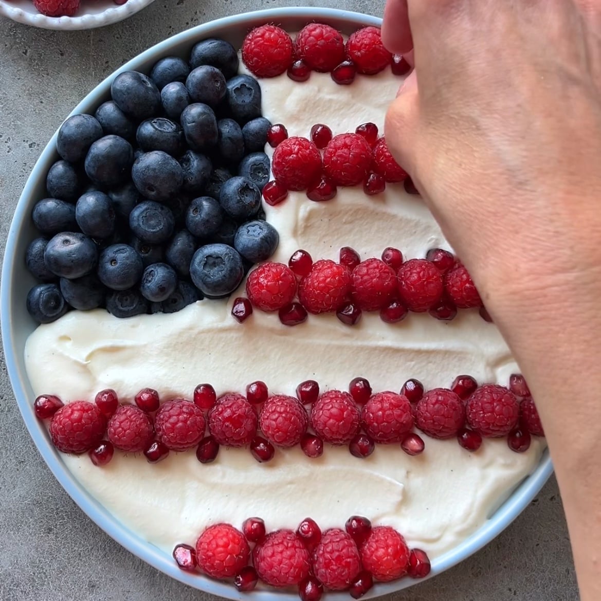 A hand decorates a dish with blueberries, raspberries, and pomegranate seeds on a creamy base, arranged to resemble the American flag.