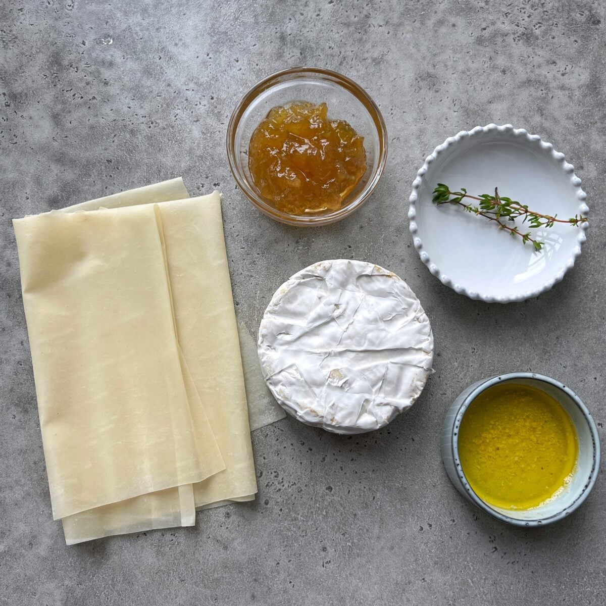 Ingredients for Baked Brie in Phyllo: phyllo dough sheets, a wheel of brie, a bowl of melted butter, fig jam, and fresh thyme sprigs.