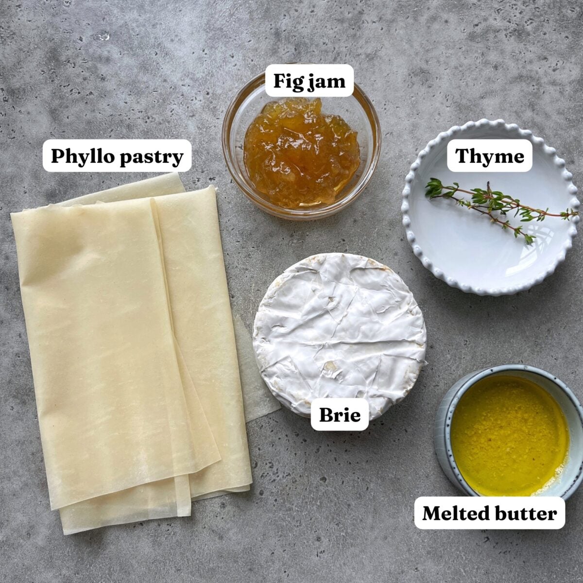 Phyllo pastry, fig jam, thyme, brie cheese, and melted butter arranged on a gray surface, each labeled with white text.