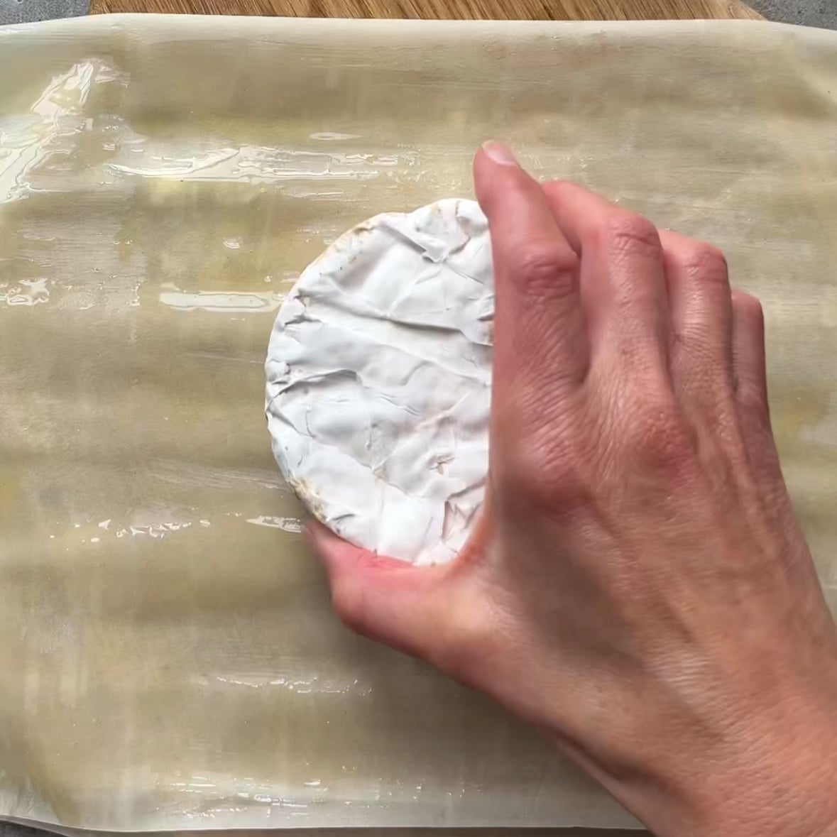 A hand places a round of cheese with a white rind onto a sheet of butter-brushed phyllo dough on a wooden surface.