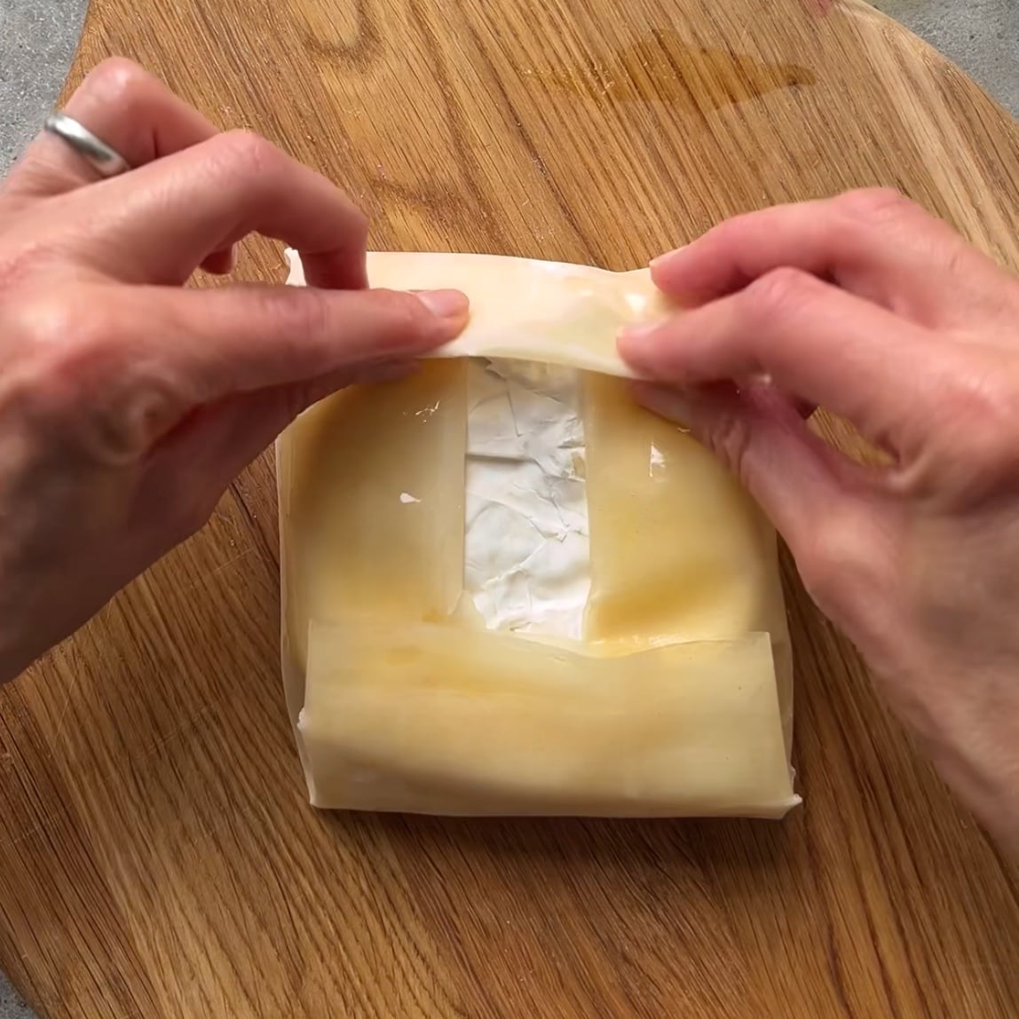 Two hands folding a sheet of pastry around a block of cheese on a wooden surface, preparing Baked Brie in Phyllo.