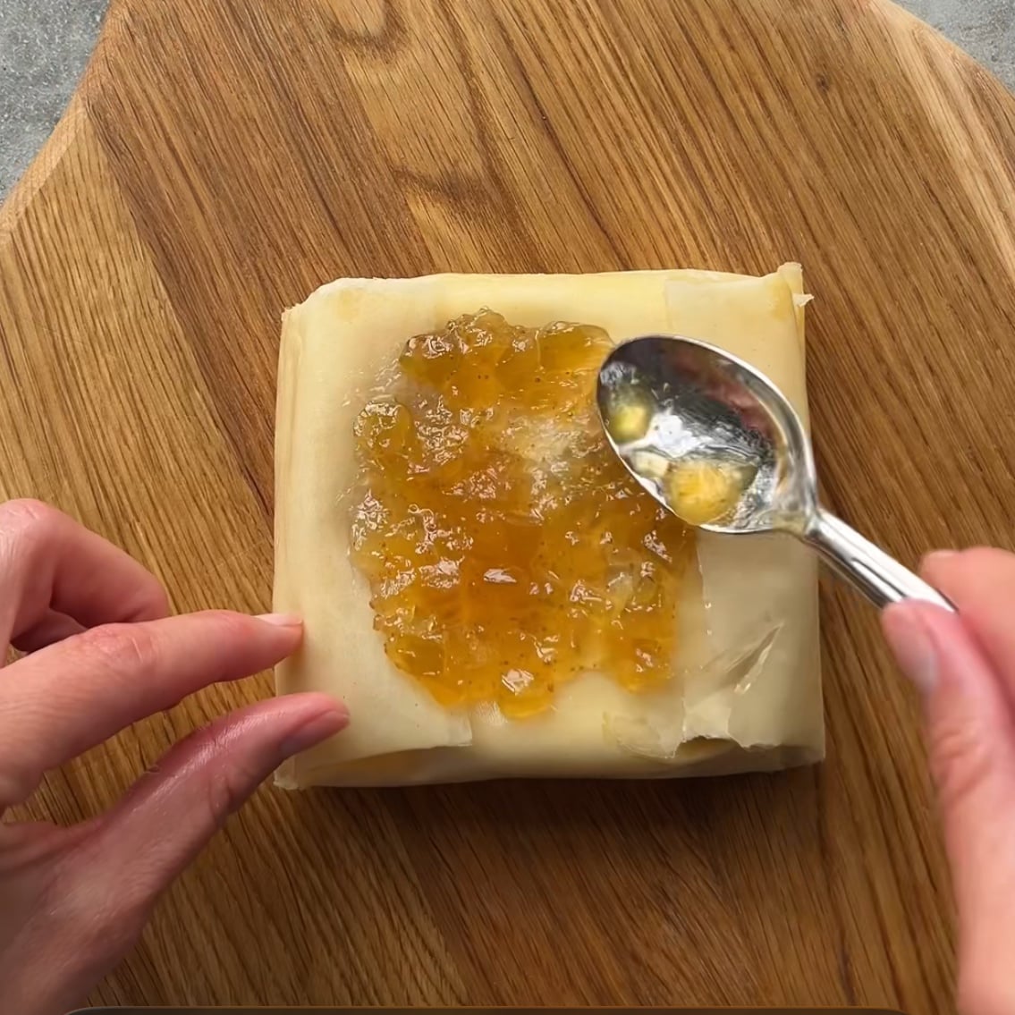 A person spreads a layer of yellow jam onto a folded pastry sheet with a spoon.