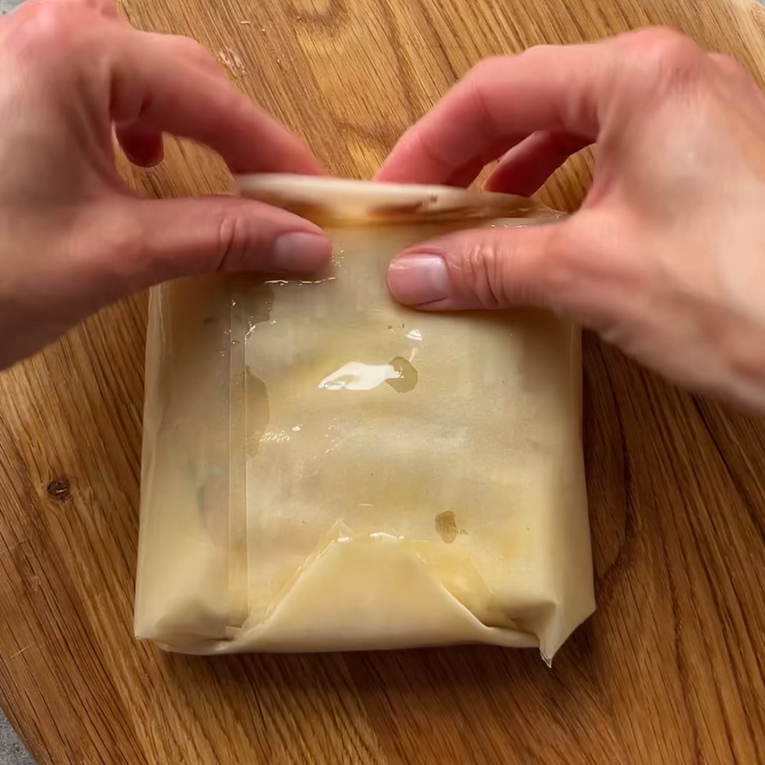 Two hands folding the top of a phyllo wrapped packet on a wooden surface, sealing in the delicious flavors.