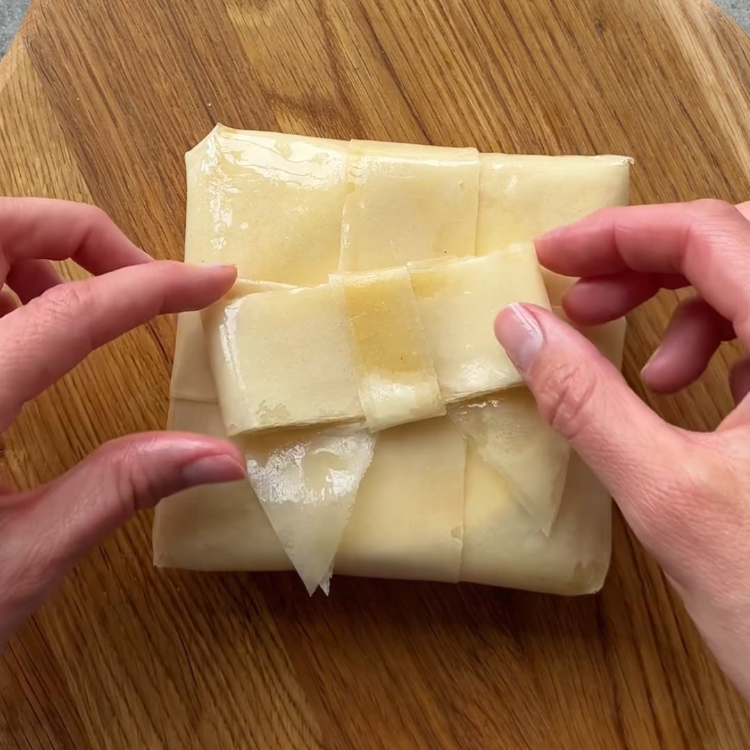 Two hands place a pastry bow on top of a rectangular pastry resting on a wooden surface.