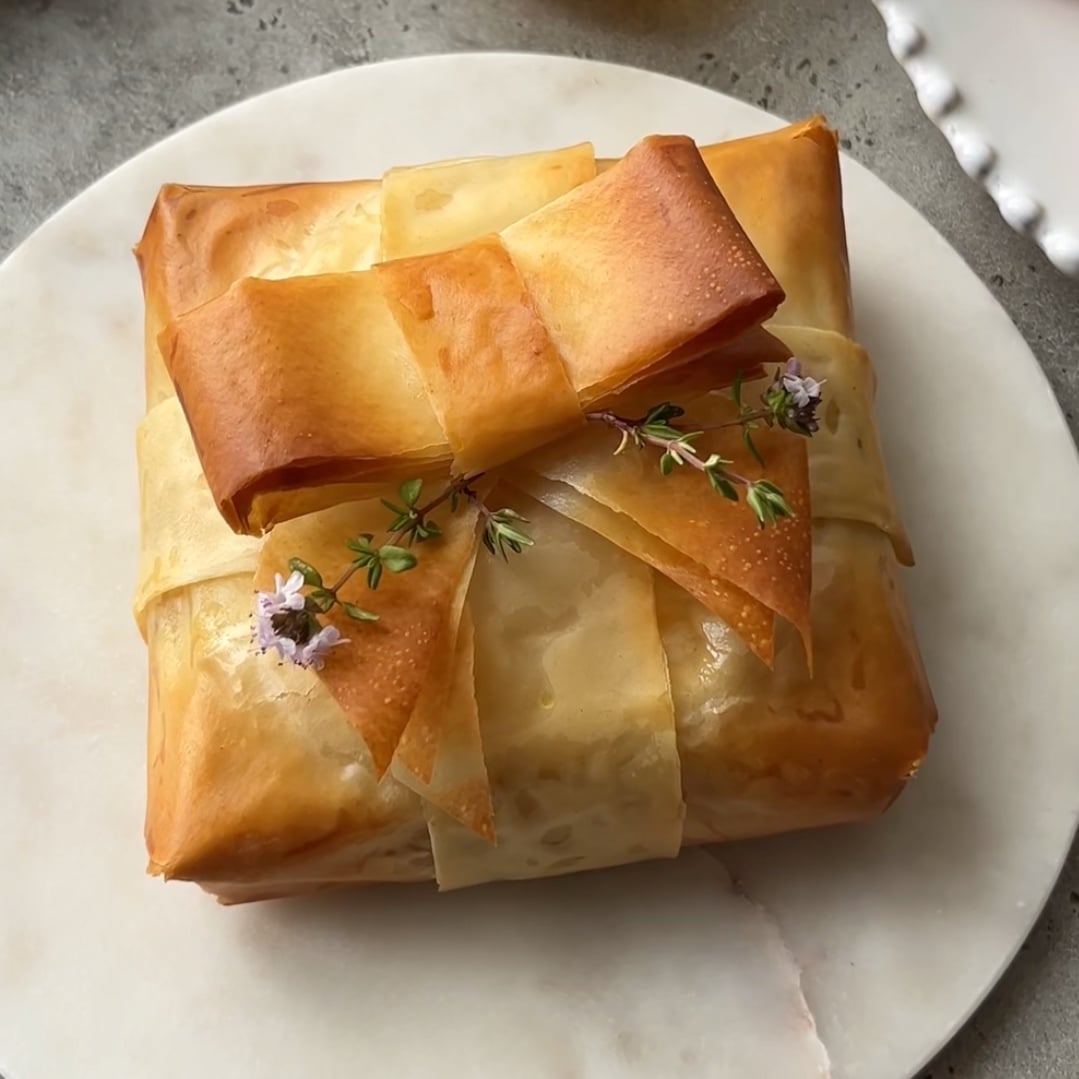 A Baked Brie in Phyllo, wrapped like a gift, is decorated with a delicate phyllo bow and small sprigs of thyme and purple flowers, presented on a round marble surface.