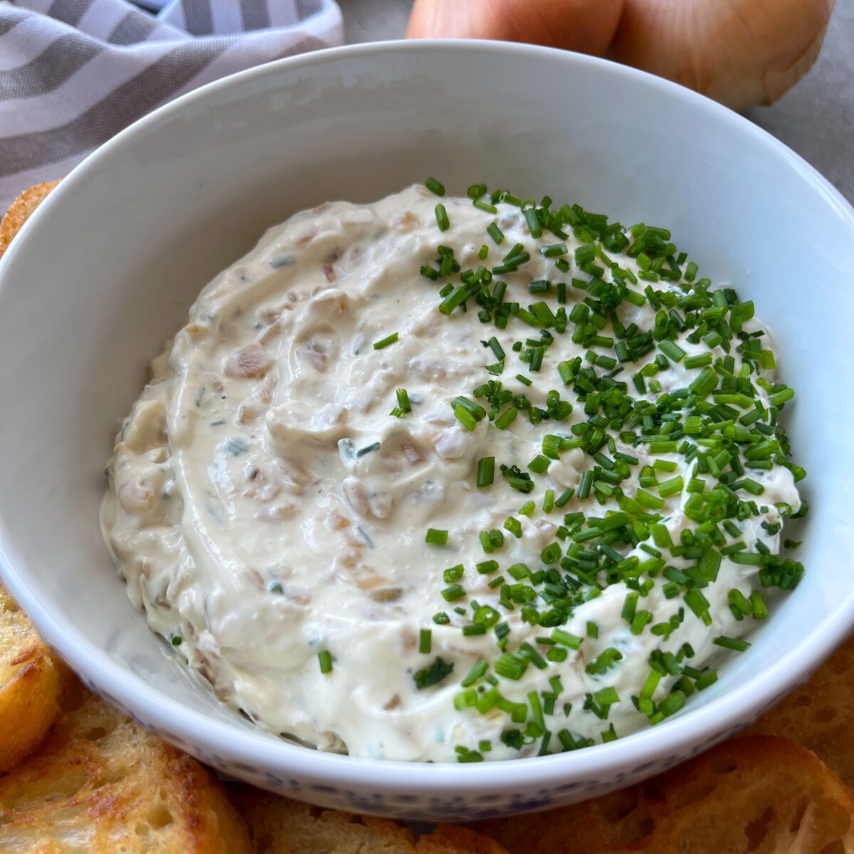 A white bowl filled with creamy caramelized onion dip garnished with chopped chives, surrounded by slices of toasted bread.