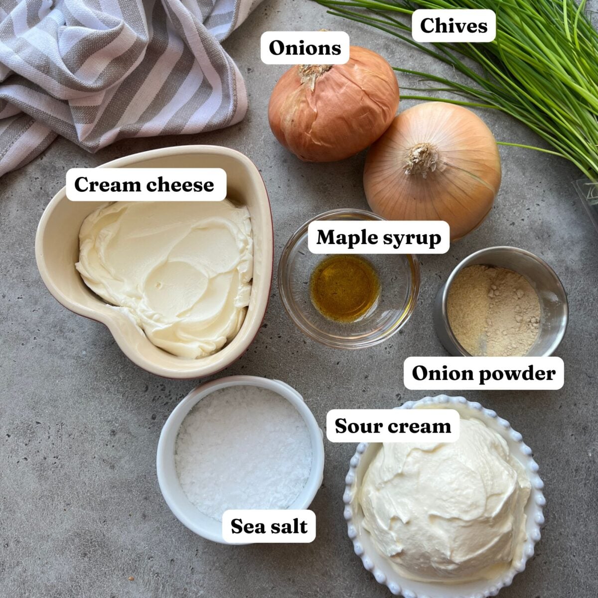 Ingredients for Caramelized Onion Dip on a counter: cream cheese, sour cream, maple syrup, onion powder, sea salt, yellow onions, and fresh chives, each labeled.