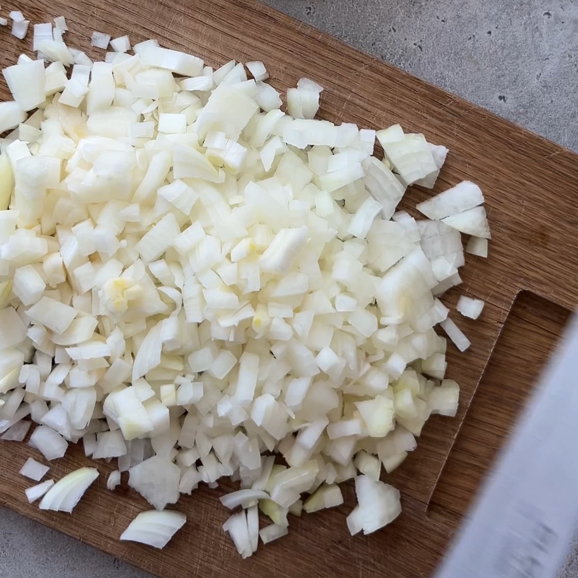 Finely chopped white onions are spread on a wooden cutting board placed on a gray countertop.