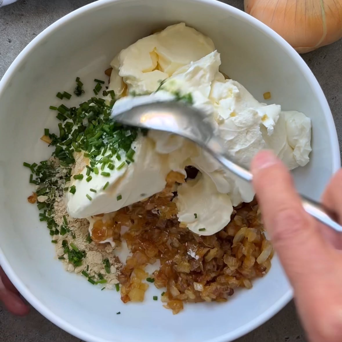 A bowl with sour cream, cream cheese, chopped chives, cooked onions, and seasonings being mixed with a spoon to create a rich and flavorful Caramelized Onion Dip.