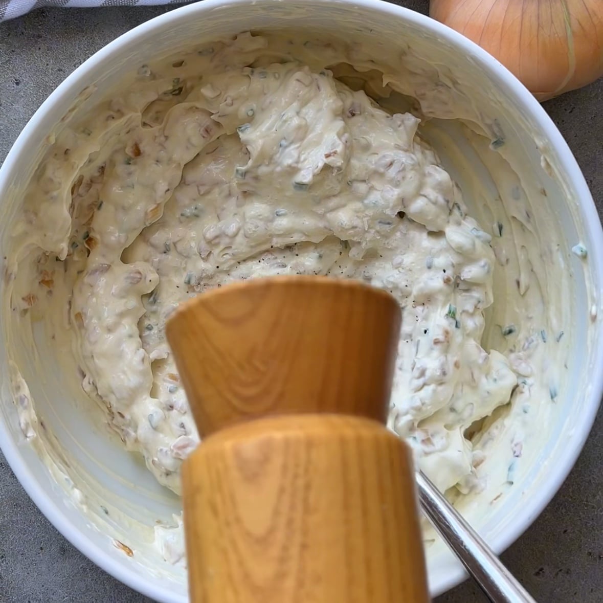 A white bowl filled with creamy Caramelized Onion Dip mixed with herbs and onions, with a wooden pepper grinder above and a spoon inside the bowl.