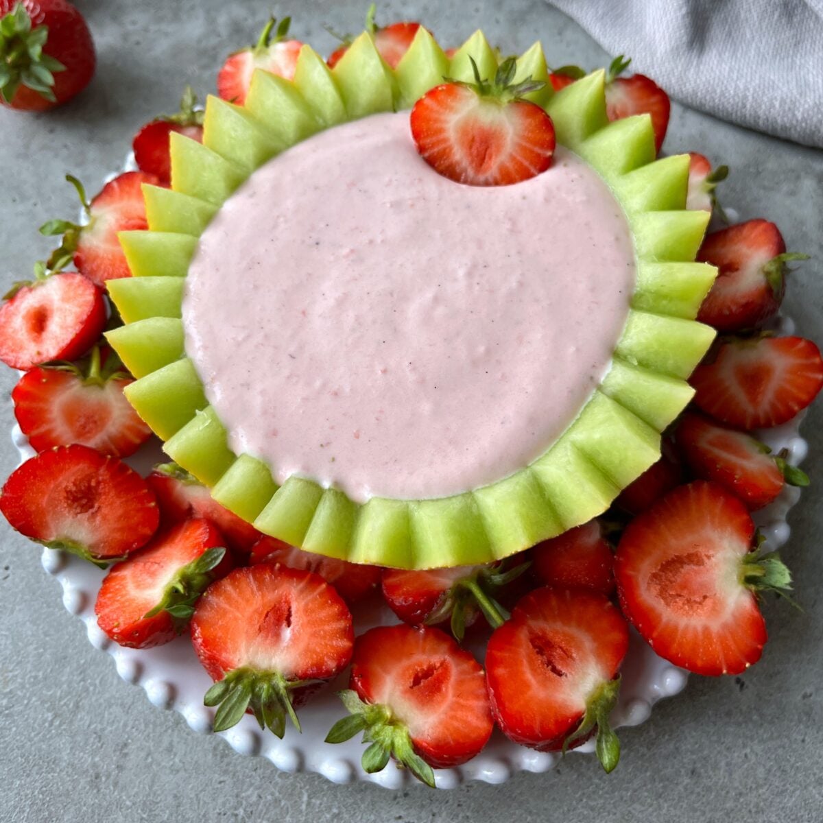 A plate of fresh strawberries surrounds a bowl made from honeydew melon, filled with creamy fruit dip.