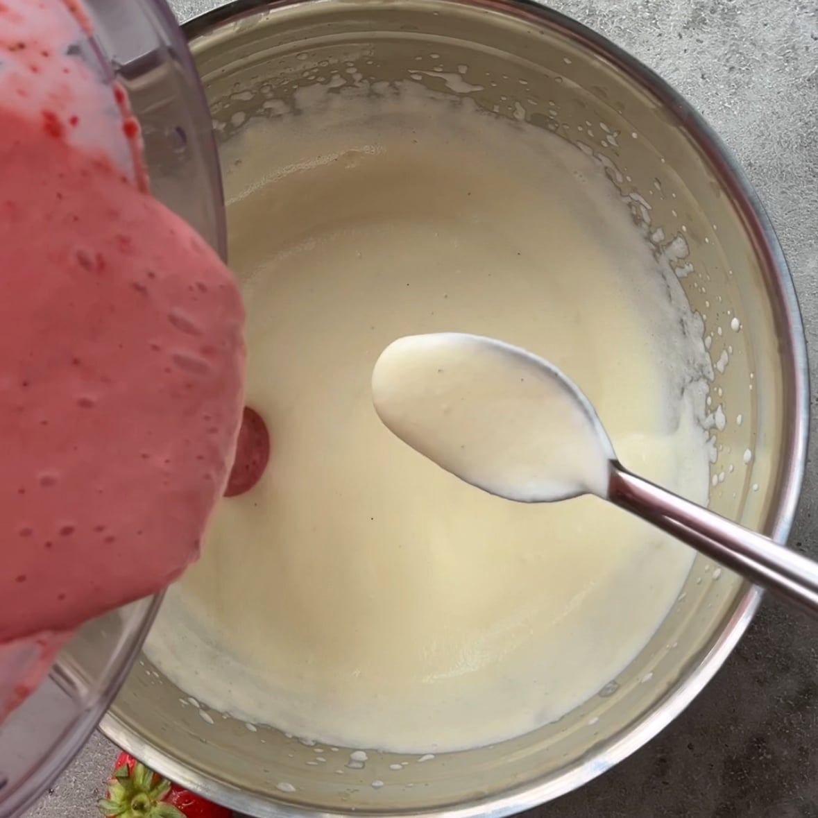 A bowl of pink strawberry mixture is being poured into a bowl of whipped cream.