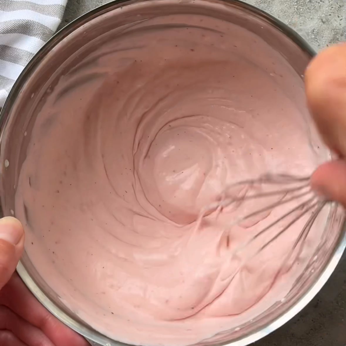 A hand whisks pink creamy dip in a stainless steel bowl on a gray surface, with a striped towel partially visible beside the bowl.