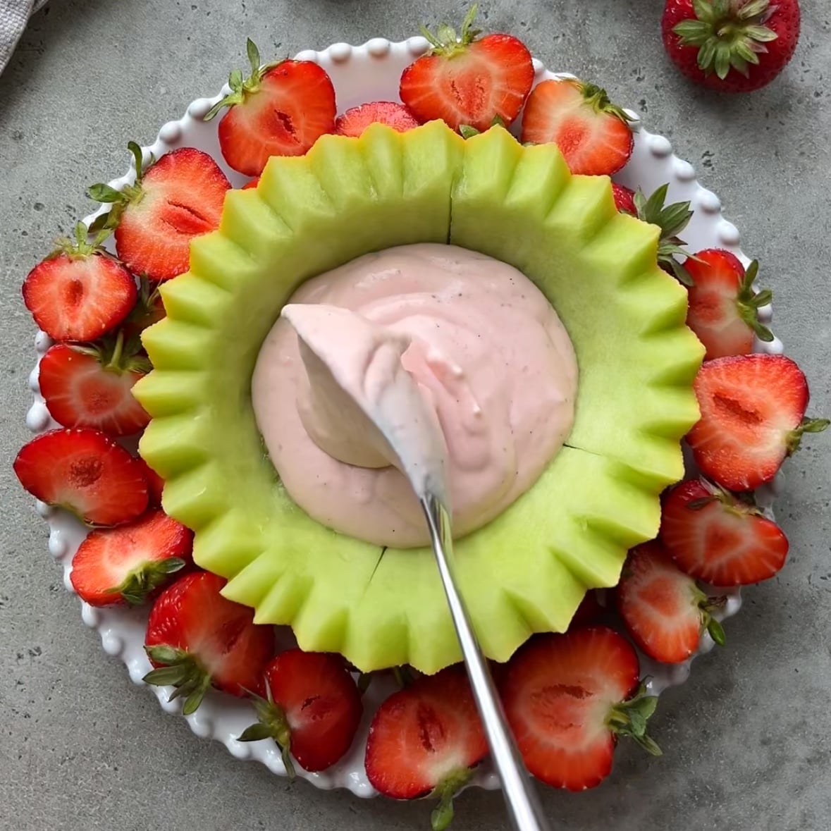 A scalloped honeydew bowl filled with pink fruit dip with cream cheese is surrounded by sliced strawberries on a white plate, with a spoon in the dip.
