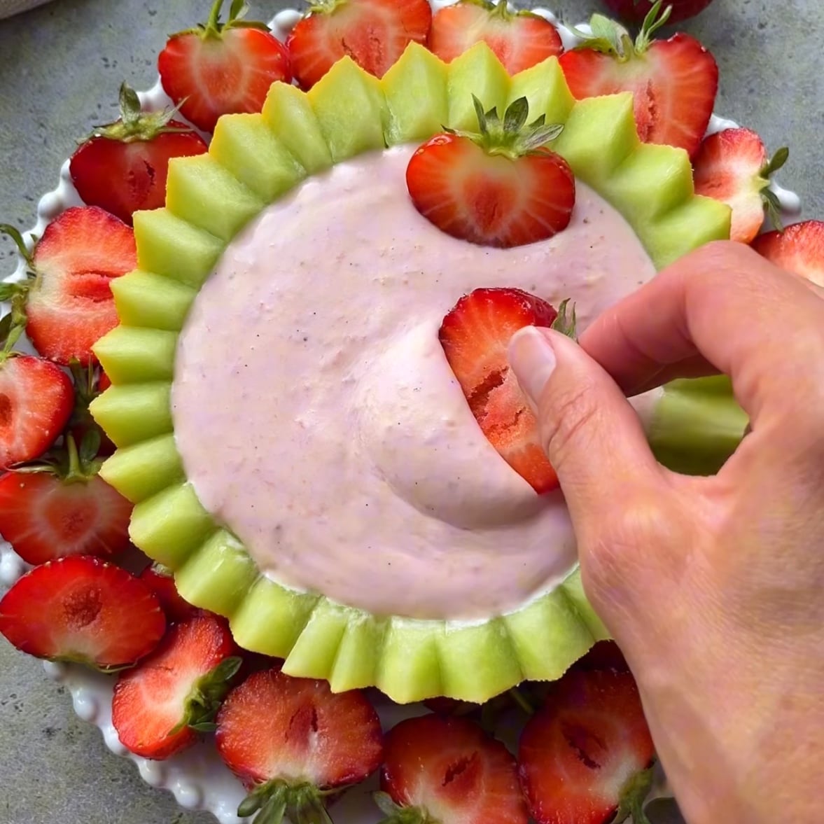 A hand dips a sliced strawberry into Fruit Dip with Cream Cheese, surrounded by more strawberries and a decorative edge of sliced melon.