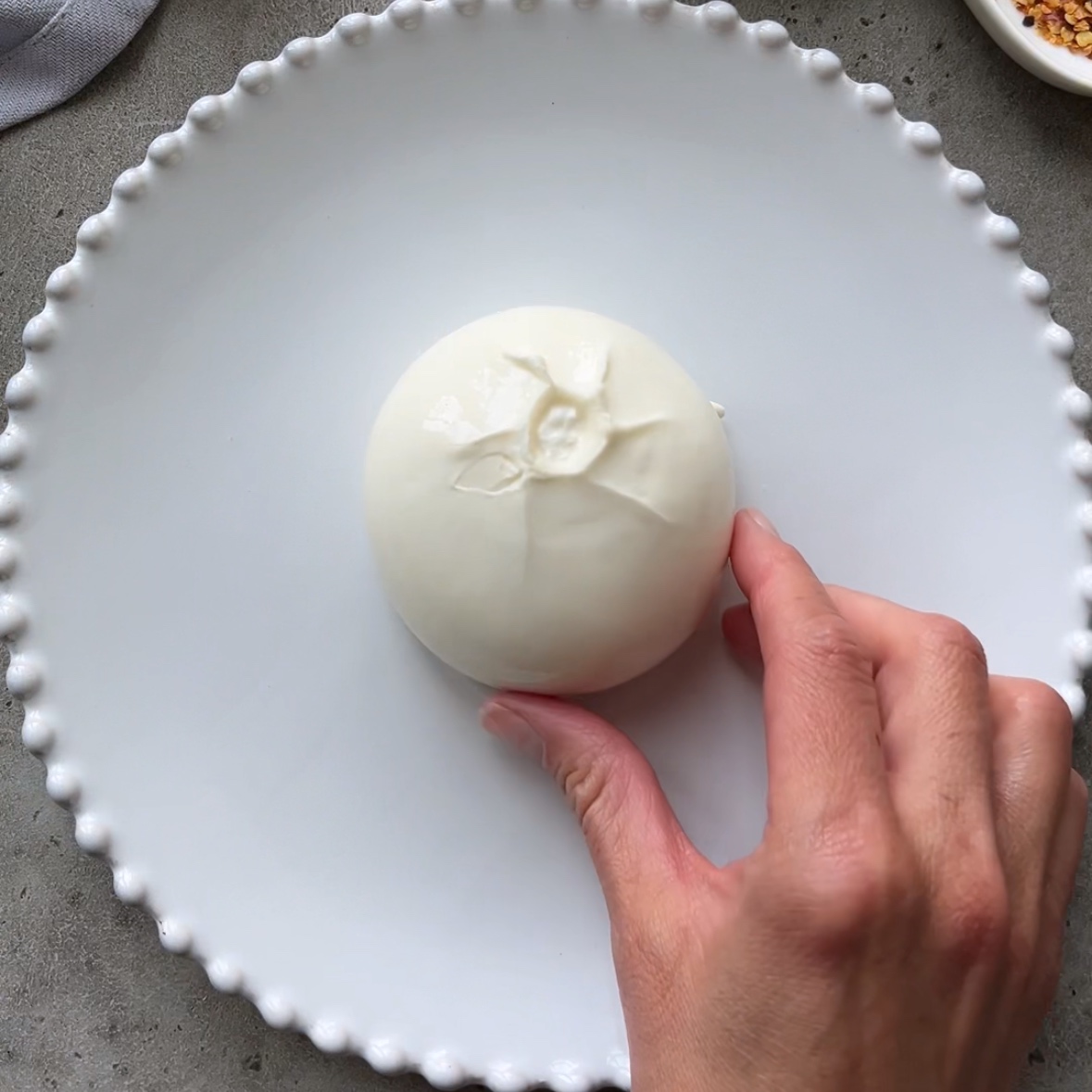 A hand places a ball of burrata cheese on a white plate with a beaded rim.